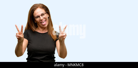 Frau mittleren Alters mit schwarzen Hemd und Brille suchen Bei Kameras Tong und Sieg mit Finger isoliert blauer Hintergrund Stockfoto