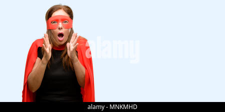 Mittleres Alter super hero Frau das Tragen der roten Maske und Cape stressig die Hände auf den Kopf, Panik, Angst, schreien blau Hintergrund isoliert Stockfoto