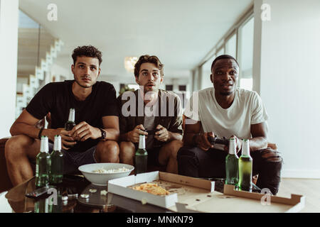 Freunde Spaß mit Getränken und Snacks beim Spielen von Spiel zu Hause. Zwei Männer spielen video game Holding Joysticks, während ein anderer Mann Uhren Esprit Stockfoto