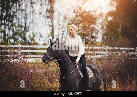 Lächelnde junge Frau in Reitbekleidung ihr Kastanie Pferd allein im Galopp durch ein Feld im Herbst Stockfoto