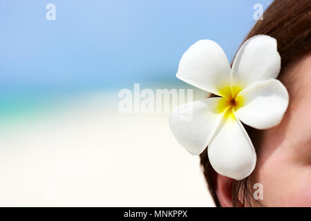 Nahaufnahme von einem weißen frangipani Blume hinter Frau Ohr mit Blick auf einen tropischen Strand Stockfoto