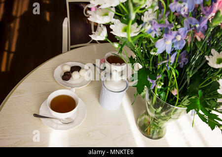 Runde weiße Tisch ist mit einem Strauß Blumen, Tassen Tee und Süßigkeiten, Sonnenlicht Stockfoto