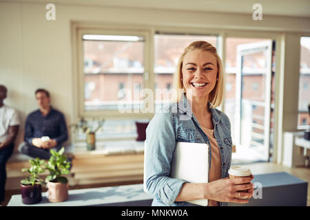 Lächelnden jungen Geschäftsfrau stehend in einem Büro mit einem Kaffee und, einen Laptop mit Kollegen im Hintergrund Stockfoto