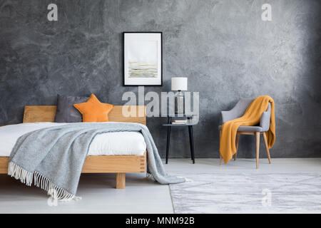 Orange Decke auf Stuhl im dunklen Schlafzimmer mit Plakat oben Lampe auf Hocker und Kingsize-Bett Stockfoto