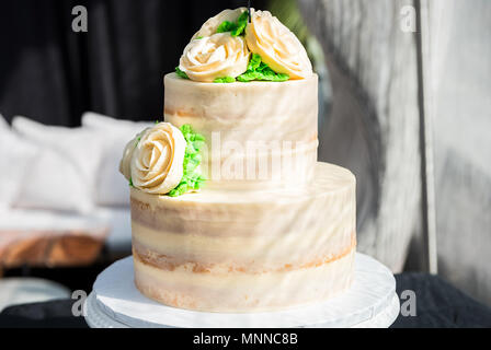 Makro Nahaufnahme von drei tiered Vanille goldenen Licht weiß gelb Creme schwamm Hochzeitstorte mit rose Blumen Dekoration auf helle, grüne Blätter Stockfoto