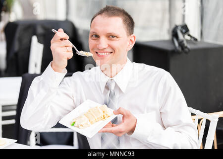 Junge glücklicher Mann im Kleid Shirt lächelnd Kuchen essen mit Gabel Hochzeit Dessert essen, sitzen am Tisch Stockfoto