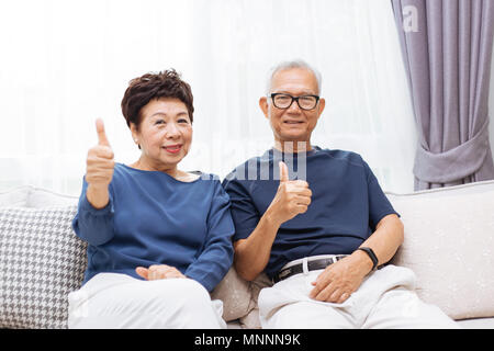 Senior asiatischen Paar an Kamera und geben Daumen bis beim Sitzen auf dem Sofa zu Hause Stockfoto