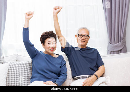 Senior asiatischen Paar an Kamera und heben die Hände auf, während er sich auf dem Sofa zu Hause sitzen Stockfoto
