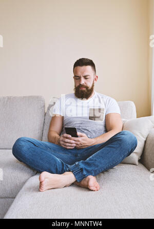 Close up Portrait von Erwachsenen kaukasischen Mann sitzt auf beige Sofa und sie ihr Smartphone Bärtigen. Internetsucht Konzept Stockfoto
