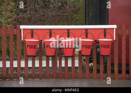 Eine Reihe von fünf red fire Eimer Wasser hängend an einem Zaun auf einem Bahnhof, die dampfzüge verwenden Stockfoto