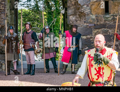 Dirleton, East Lothian, Schottland, UK, 19. Mai 2018. Dirleton, East Lothian, Schottland, UK, 19. Mai 2018. Dirleton Castle Angriff Re-enactment. Die historische Saltire Society staging a tongue-in-cheek Re-enactment eines Angriffs von Robert the Bruce, König von Schottland, um 1311, das Schloss aus dem Englischen, in authentischer Kleidung und Rüstung gekleidet und schwingende replica Waffen im historischen Umfeld Schottland Dirleton Castle. Ein Mann spielt Robert Bruce gekleidet im Kostüm mit einem Löwen in der Menge mit einem Mikrofon. Soldaten mit Spießen werden im Hintergrund Stockfoto