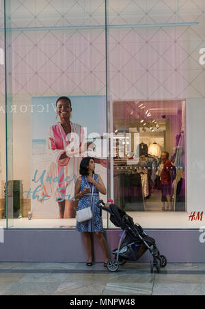 Eine junge Frau mit einem Baby im Kinderwagen stehend außerhalb der Frauen kleidung shop in einem Einkaufszentrum zu einem Mobiltelefon oder tragbares Gerät suchen. Stockfoto