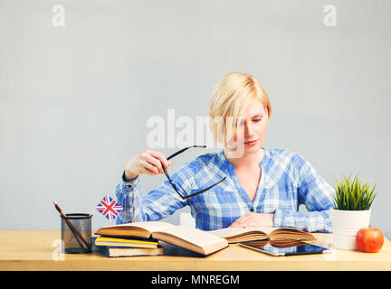 Smart Studentin, blonde Frau hält eine Brille in der Hand, Studium der Englischen Sprache auf Tisch mit Büchern, erfolgreiches Lernen Konzept Stockfoto
