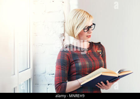 Ziemlich intelligente blonde Frau trägt Brillen und roten Kleid lesen Buch in der Nähe von weiten Fenster in laght Zimmer Stockfoto