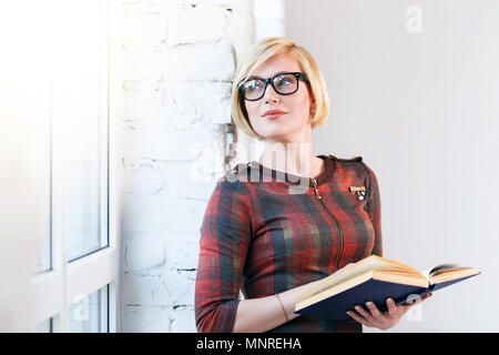 Ziemlich intelligente blonde Frau trägt Brillen und roten Kleid lesen Buch in der Nähe von weiten Fenster in laght Zimmer Stockfoto