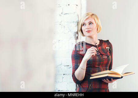Ziemlich intelligente blonde Frau trägt Brillen und roten Kleid lesen Buch in der Nähe von weiten Fenster in laght Zimmer Stockfoto