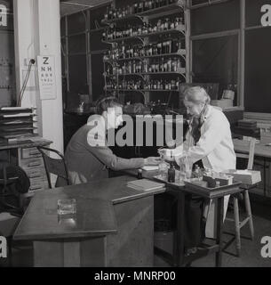 1950, historische, eine junge Frau, die ihre Hand von einer älteren Frau Doktor an ein Health Centre, London, England, UK behandelt. Beachten Sie die große Auswahl an verschiedenen flüssigen Arzneimitteln in Glasflaschen auf Regalen in der Chirurgie und das Auge Test Board an der Wand hängen. Stockfoto