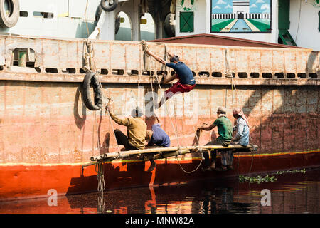 Dhaka, Bangladesh, 24. Februar 2017: Maler auf einer Werft in Dhaka sitzen auf einem Holzbrett und tragen auf dem Rumpf eines Schiffes auf dem Rostschutz Stockfoto