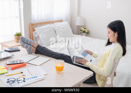 Asiatische Frau Freiberufler lesen Bericht Papier und Arbeiten mit Laptop lag auf Knie mit Rest Bein am Tisch im Schlafzimmer zu Hause. Arbeit zu Hause Konzept. Arbeit f Stockfoto