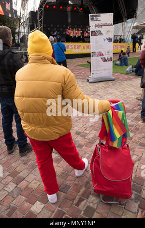Frau gekleidet in gelben Hut gold Jacke und rote Hosen hält ein rotes shopping Trolly und bunte Fahne, eine musikalische Leistung. Stockfoto