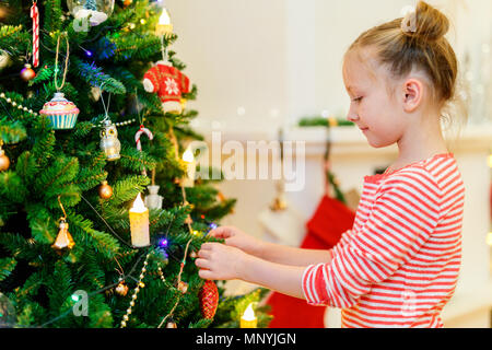 Adorable kleine Mädchen schmücken Weihnachtsbaum mit bunten Christbaumkugeln aus Glas zu Hause Stockfoto