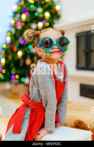 Adorable kleine Mädchen mit lustig Weihnachten Gläser in Home schön mit Weihnachtsbaum und Lichtern geschmückt Stockfoto