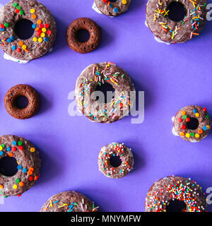 Schokolade gebackene Donuts auf einem violetten Hintergrund verglast und mit Konfetti eingerichtet. Ansicht von oben. Quadratisches Bild. Stockfoto