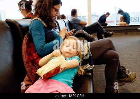 Eine Mutter mit ihrem iphone, während ihre Tochter entspannt, lehnte sich zurück auf ihre Oberschenkel essen eine Tüte Chips beim Warten auf ihren Flug aufgerufen werden Stockfoto