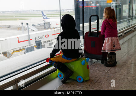 Zwei Kinder (5 und 3 Jahre alt, Bruder und Schwester) mit ihren Koffer heraus aufpassen durch das Fenster am Flughafen London Heathrow Stockfoto