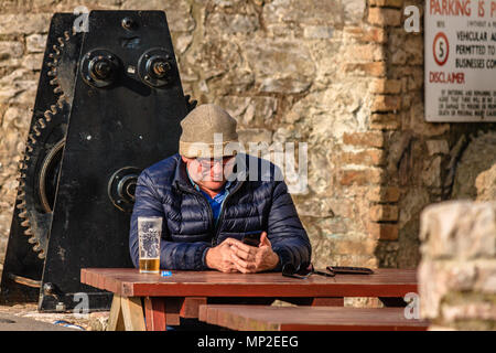 Mann in warmen Mantel und Hut saßen draußen ein Pub mit einer fast fertigen Pint Bier und über ein Mobiltelefon. Teignmouth, Devon. Feb 2018. Stockfoto