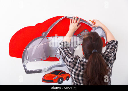 Eine Nahaufnahme einer weiblichen Künstlerin zeichnet ein Bild von einem Auto und zieht es in Rot auf einer weißen Wand im Kinderzimmer Stockfoto