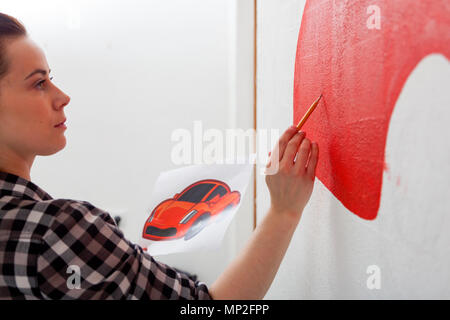 Nahaufnahme einer Künstlerin in schwarzen und weißen Hemd zeichnet ein rotes Auto an der Wand mit einem Bleistift auf eine weiße Wand im Kinderzimmer Stockfoto