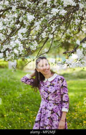 Schöne brünette Frauen tragen hübsches Kleid mit Blumendruck in der Nähe von blühender Apfelbaum Stockfoto