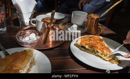 Hand schneiden einen Teil der spanakopita, eine traditionelle griechische Spinatkuchen auf eine Tabelle mit Kupfer Kanne mit Kaffee, im Freien. Stockfoto