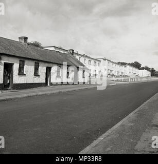 Ein historisches Bild der 1950er Jahre, das eine Reihe von traditionellen, aus Stein gebauten einstöckigen Hütten zeigt und dann weiter die Straße hinauf, aber etwas zurückgelegter eine Reihe von neu gebauten, modernen, zweistöckigen Häusern, Irland. Stockfoto