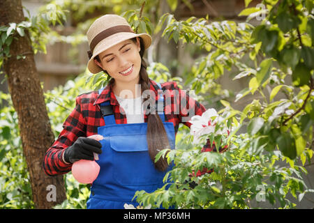 Glückliche junge Frau Gärtner in Arbeit Kleidung Sprühen von Pestiziden oder Wasser auf ihr Pfingstrose Blüten. Gartenbau und Pflege der Pflanzen als Hobby Konzept Stockfoto