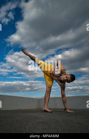 Austrickst, auf die Straße. Kampfsportarten Mann macht Kick mit Support für Arm und Bein barfuß. Bhudda Verkürzungen von unten gegen den Himmel. Stockfoto