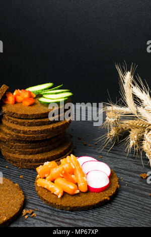 Scheibe Schwarzbrot mit einem Schnitt in der Form eines Herzens mit Gemüse - Gurken, Radieschen, rote Paprika, Karotte auf dunklem Hintergrund. Gesundheit Stockfoto