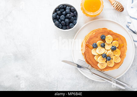 Buttermilch Pfannkuchen mit Banane, Blaubeeren und Honig auf konkreten Hintergrund. Ansicht von oben und kopieren Sie Platz für Text. Blick von oben auf die Pfannkuchen mit Beeren auf Stockfoto