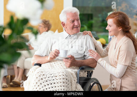 Happy gelähmt älterer Mann im Rollstuhl Tee trinken und ein Betreuer die ihn unterstützen Stockfoto