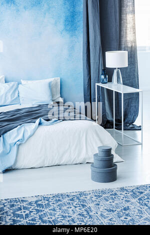 Gemusterten Teppich und grauen Kästchen neben dem Bett mit blau Bettwäsche in minimalen Schlafzimmer Innenraum mit Lampe Stockfoto