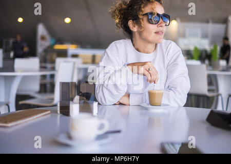 Schönen kaukasischen Frauen mittleren Alters mit Sonnenbrille, und trinken Sie einen Kaffee in einer Bar. outdoor Licht vom Fenster für eine Freizeit rest Moment während des Stockfoto