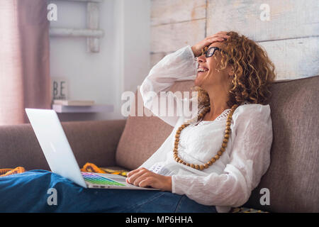 Lächeln und Lachen kaukasischen schönen mittleren Alter Frau, sitzen Sie auf dem Sofa zu Hause mit einem farbigen Tastatur Laptop. Arbeiten gerne unabhängige Konzept Stockfoto