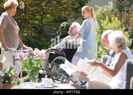 Krankenschwester Unterstützung älterer Mann im Rollstuhl beim Treffen mit Freunden im Garten an einem sonnigen Tag Stockfoto