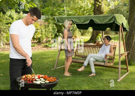 Aufnahme eines jungen Mannes grillen Essen und seine Freunde sprechen im Hintergrund Stockfoto