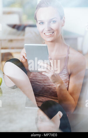 Die moderne Frau in Sportkleidung mit Tablette beim Sitzen auf Stuhl in der Küche Stockfoto