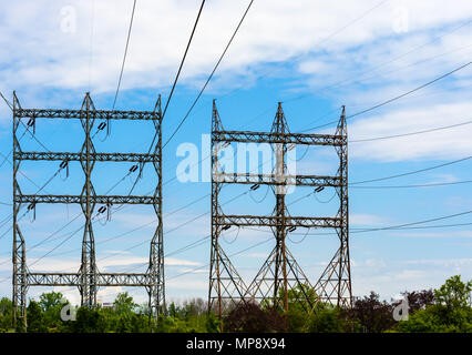 Zwei große rechteckige elektrische Sendemasten mit Stromleitungen und Isolatoren über Bäumen unter teilweise bewölktem Himmel. Stockfoto