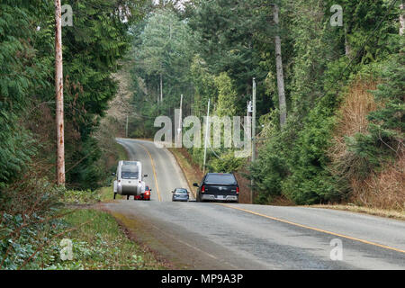 Auf eine Landstraße durch den Wald, drei Autos fahren einen Hügel hinunter, während ein 97 Jahre alter Mann ein elektrisches Dreirad den Hügel hinauf, der von einem Auto verfolgt Pedale. Stockfoto