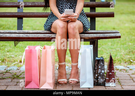 Nahaufnahme einer Frau Beine sitzen auf der Bank im Park und Surfen auf einem Smartphone mit Tüten neben. Stockfoto
