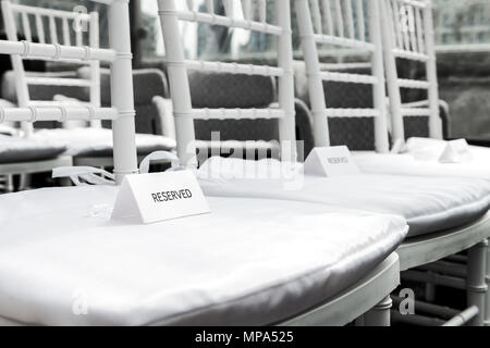 Nahaufnahme des White Wedding Stühle mit reservierten Zeichen für die Zeremonie mit Hintergrund der Zeilen von viele Sitze Muster, niemand Stockfoto